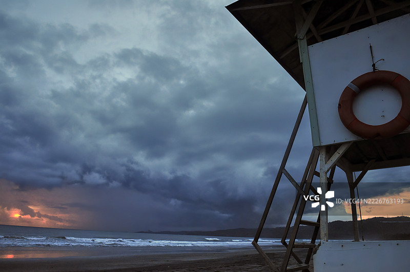 一个白色的救生员小屋，旁边有一个红色的救生圈和旗帜，矗立在黑暗的海滩上。图片素材