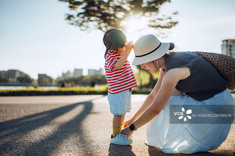 一位关心的年轻亚洲母亲在公园里帮助并教她的小女孩系鞋带。家庭的爱、关心和支持的理念。图片素材