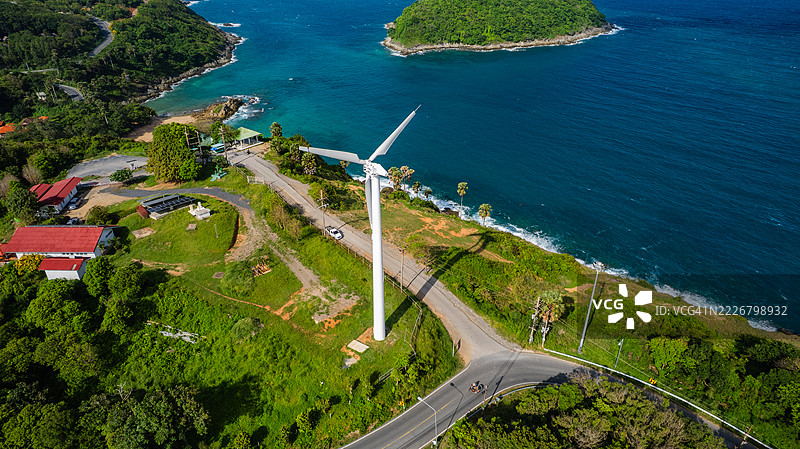 海岸风力涡轮机的无人机照片，周围环绕着绿色植被、蓝色海洋、蜿蜒的道路和岛屿风景，宣传可再生能源。图片素材