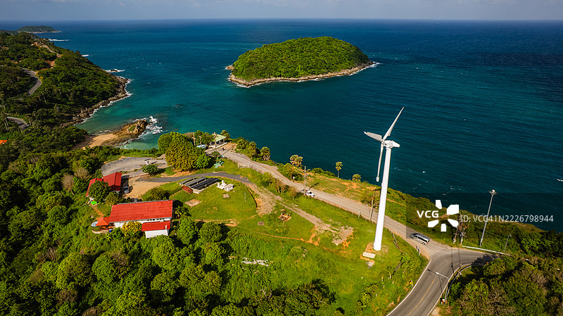 海岸风力涡轮机的无人机照片，周围环绕着绿色植被、蓝色海洋、蜿蜒的道路和岛屿风景，宣传可再生能源。图片素材