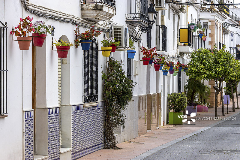 西班牙安达卢西亚马拉加省埃斯特波纳的老城区卡斯科安蒂瓜的一条街道,白色房屋墙壁上挂着几朵花卉糖果。图片素材