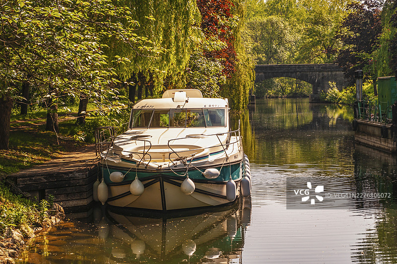 在树木成荫的河流上，房船悠然航行，背景是石桥，位于法国新阿基坦大区的沙朗特省。图片素材
