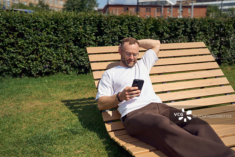 放松的男人在绿色公园的木质躺椅上悠闲地坐着，微笑着在阳光明媚的日子里浏览智能手机。图片素材