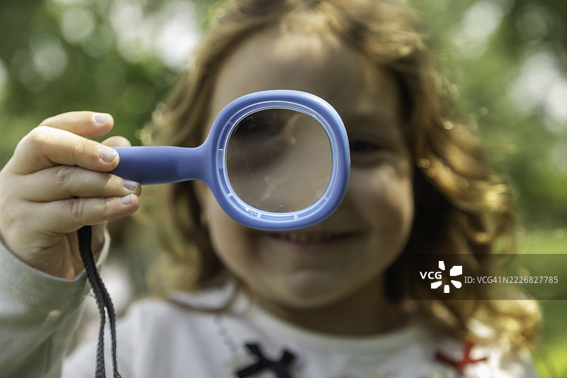阳光明媚的公园里，一个微笑的小女孩手持放大镜的肖像图片素材