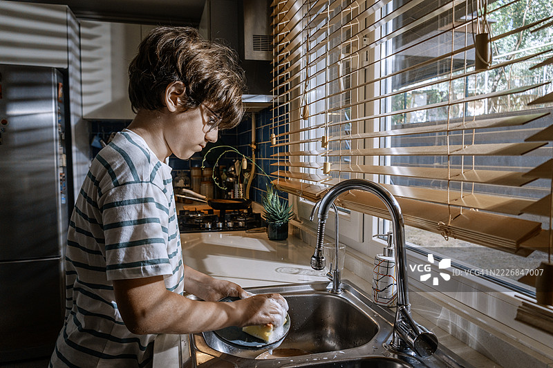 男孩在厨房水槽里洗碗，窗帘是百叶窗。图片素材