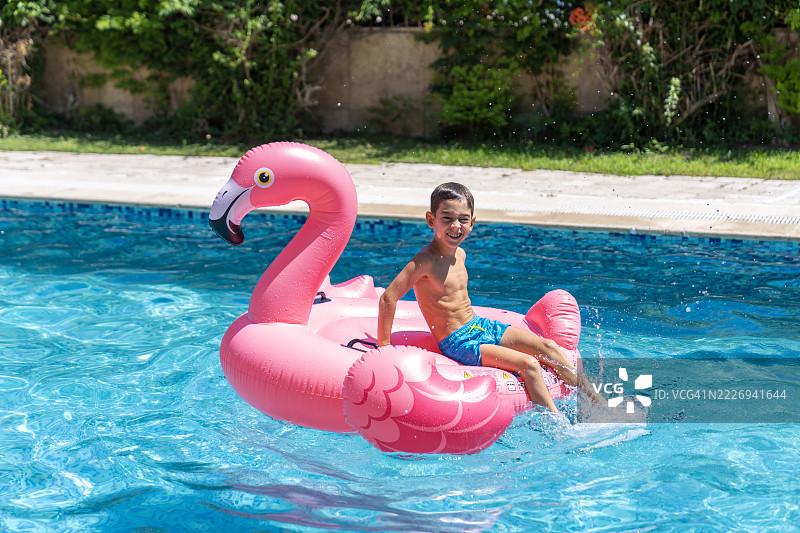 年轻男孩坐在粉色火烈鸟充气浮筒上。年轻男孩在游泳池里与粉色火烈鸟玩耍。图片素材