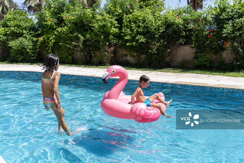 孩子们坐在粉色火烈鸟充气浮筒上。孩子们在游泳池里与粉色火烈鸟玩耍。图片素材