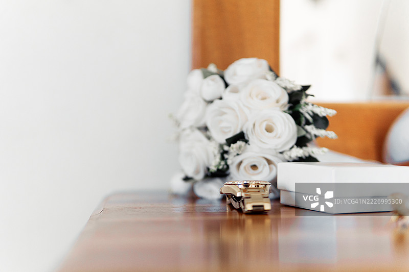 精致的白玫瑰、金色怀表、珍珠首饰静静地摆放在风化的木质梳妆台上，柔和的灯光突显了新娘早晨准备的细节。图片素材