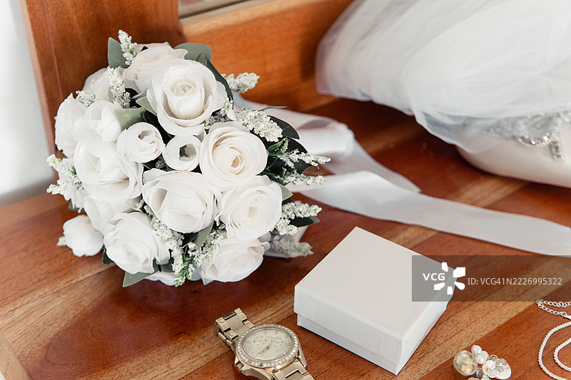 白色玫瑰花束静静地放在精致的婚礼配饰旁边 - 珍珠耳环、银项链、手表 - 背景是柔和的婚纱，衬托在质朴的木质表面上图片素材