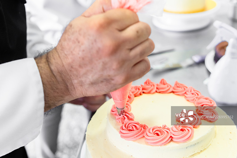糕点师在专业厨房中小心地从裱花袋中挤出粉色糖霜，制作精致的玫瑰花装饰蛋糕。图片素材