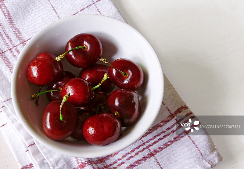 一只白色碗里装着红樱桃，放在红白相间的餐巾上。背景为白色。图片素材