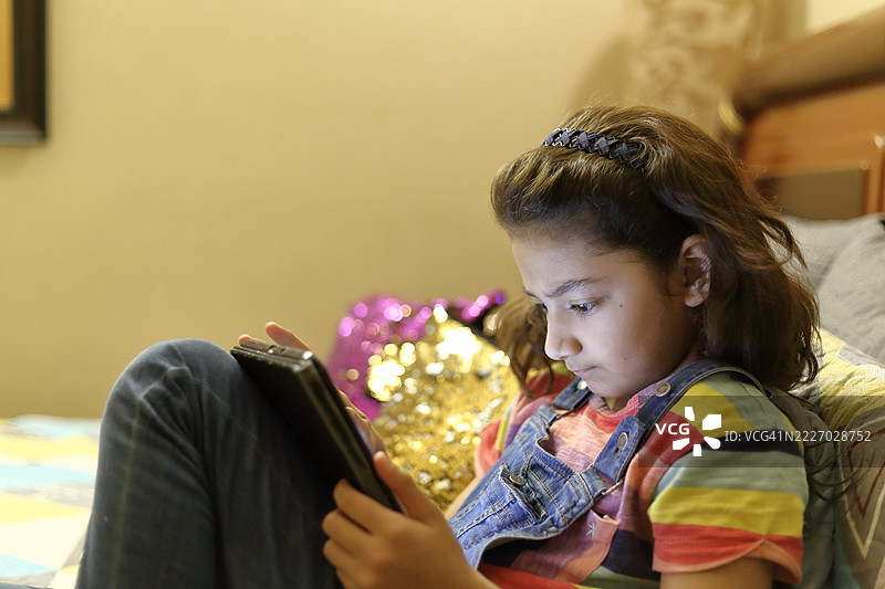 一个年轻的亚洲女孩在家卧室的床上坐着，晚上使用数字平板或手机。图片素材