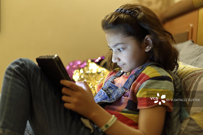 一个年轻的亚洲女孩在晚上微笑着坐在家里的床上，使用数字平板或手机。图片素材