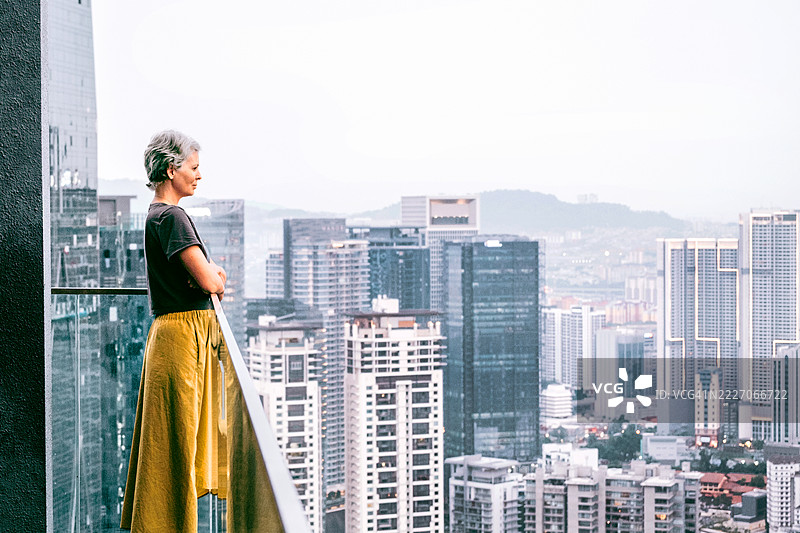 美丽的白人女性短发灰色，双臂交叉站在阳台上，凝视着高楼大厦。城市天际线景观。图片素材