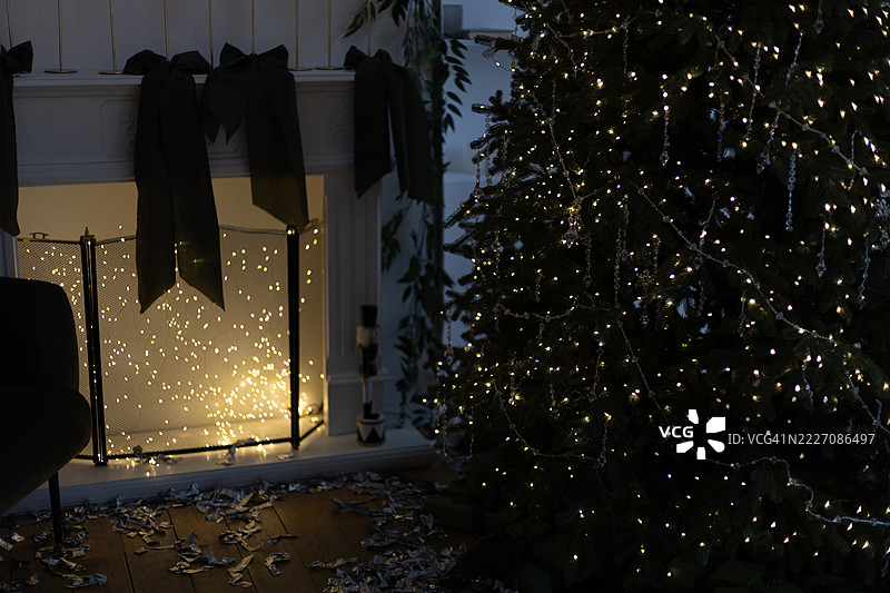 圣诞树上挂满闪烁的灯饰和装饰品，旁边是一个装满发光灯饰的壁炉屏风，地板上铺着亮片，整个房间显得昏暗。图片素材