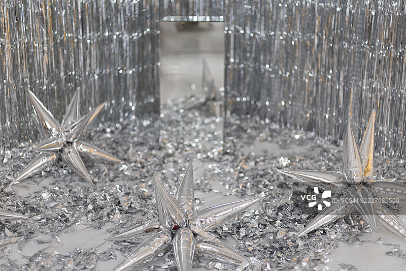 大型银色星形气球散落在覆盖着金属彩纸的地板上，背景是银色的闪光流苏。图片素材