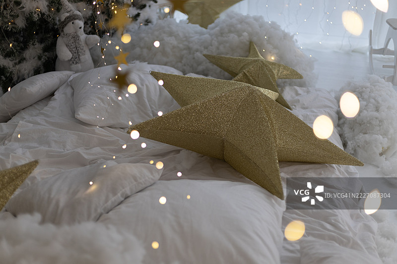 舒适的床铺，配有白色床上用品和枕头，大型金色星星装饰，蓬松的云朵元素，圣诞树旁的小雪人，所有这些都被散景灯光照亮。图片素材