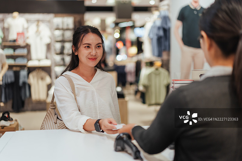亚洲女性在服装店向收银员递交信用卡，微笑着进行支付，背景是衣物货架——展现积极的购物和客户服务互动。图片素材