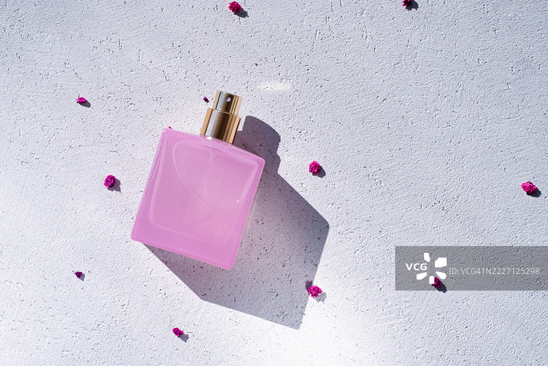 优雅的粉色香水瓶放置在有纹理的表面上，周围散落着花瓣，阳光明媚。图片素材