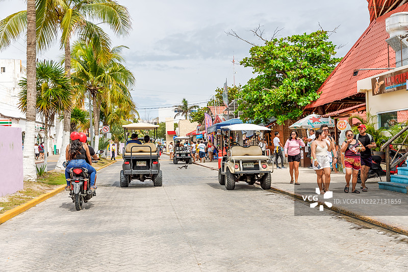 墨西哥伊斯拉穆赫雷斯岛上，繁忙的街道上人们悠闲地漫步，周围有高尔夫球车、商店和餐馆，靠近渡轮。图片素材