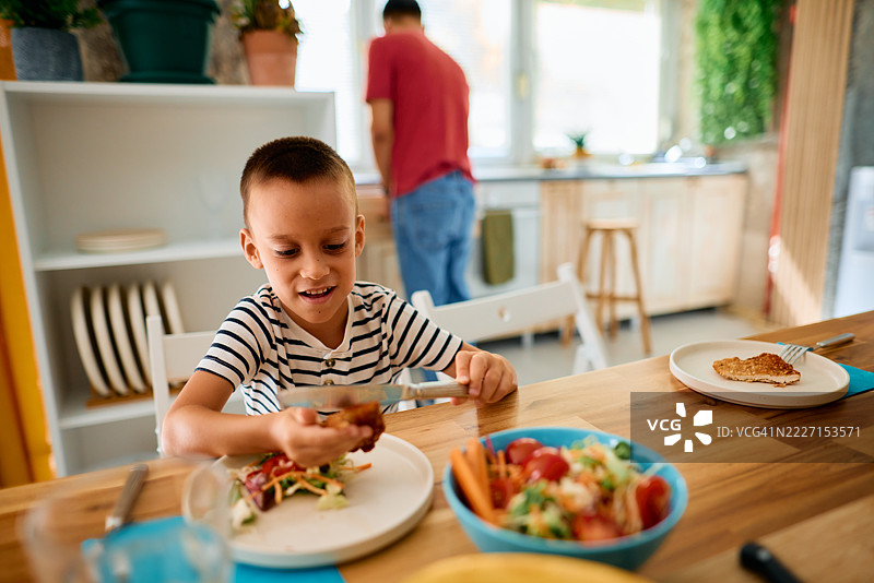 快乐的男孩在家里享用健康餐，父亲在背景中准备食物图片素材