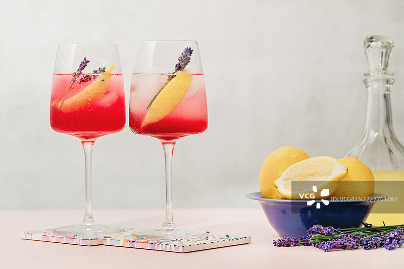 夏季花园桌上摆放着两杯薰衣草柠檬气泡鸡尾酒，旁边有一瓶柠檬酒，酒杯上有水珠。图片素材