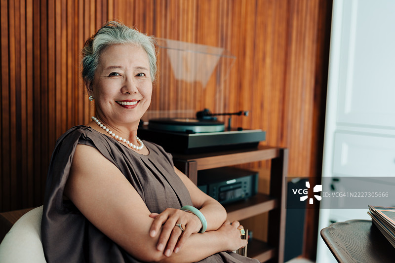 亚洲老年女性在家中客厅享受唱片，面带微笑地看着镜头。轻松的生活方式，人与退休的概念。图片素材