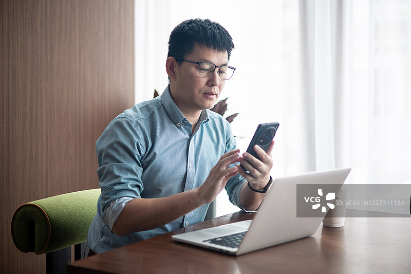 商人在办公室使用笔记本电脑工作，同时用手机发短信或使用社交媒体。图片素材