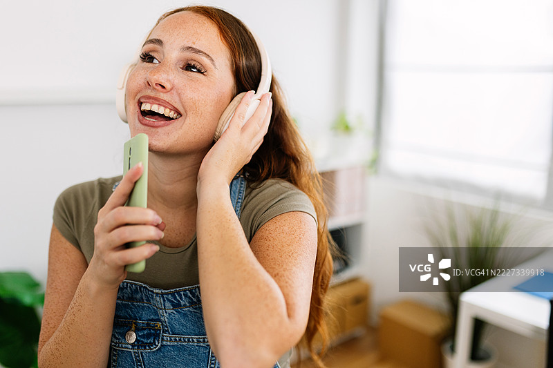 年轻快乐的女性在家中戴着耳机和智能手机唱歌图片素材