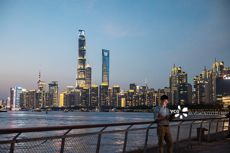 年轻的亚洲商人在上海金融区的夜晚，明亮的企业摩天大楼前使用数字平板电脑。图片素材