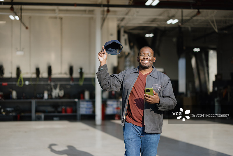 快乐的机械师在车库里挥舞着帽子并使用智能手机图片素材