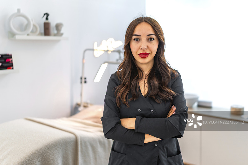 自信的女美容师双臂交叉站立在现代美容沙龙中，代表着美容行业的专业和专长图片素材