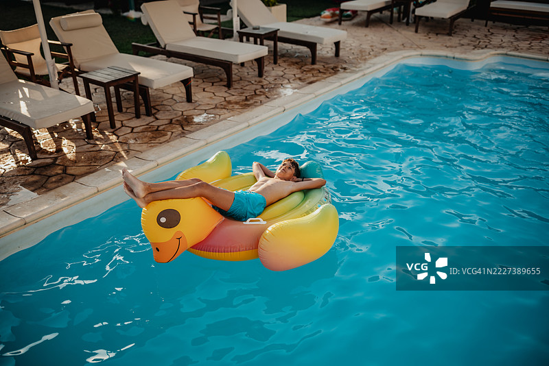 小男孩在阳光明媚的日子里，坐在五彩斑斓的充气鸭子上享受游泳池的乐趣。图片素材