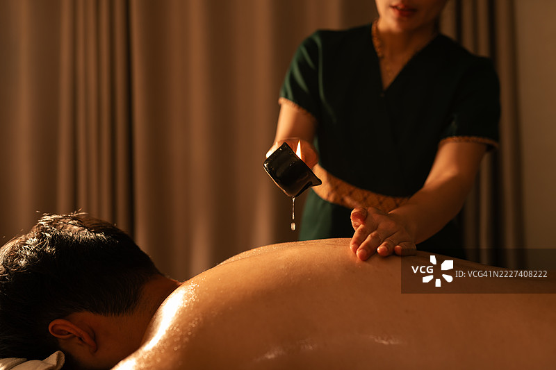 泰国治疗师将融化的按摩蜡烛中的温暖油倒在一名男子的背上，营造出宁静柔和的健康环境中的舒缓和治疗水疗体验。图片素材