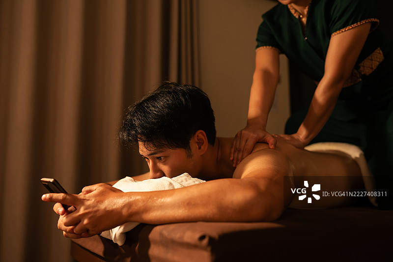 亚洲男子在使用智能手机时接受传统泰式按摩。现代健康理念展示了数字干扰与放松水疗环境中身体疗法之间的对比。图片素材