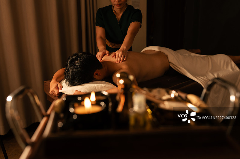 亚洲男子在昏暗的水疗室享受放松的背部按摩。专业治疗师进行传统泰式疗法。香薰蜡烛和精油增强了宁静的氛围。图片素材