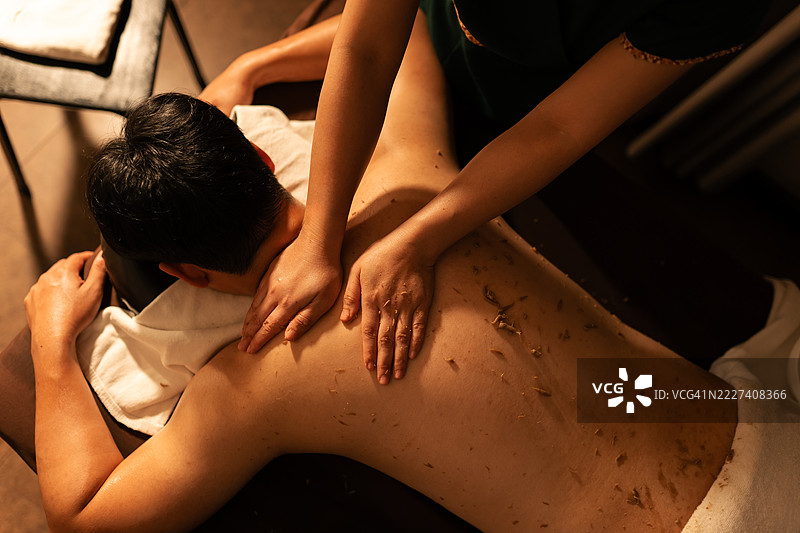 从上方俯视，治疗师在水疗中心为亚洲男性的背部涂抹草本身体磨砂膏。可见天然去角质剂和治疗性的手部动作。男性健康、护肤和传统身体护理。图片素材
