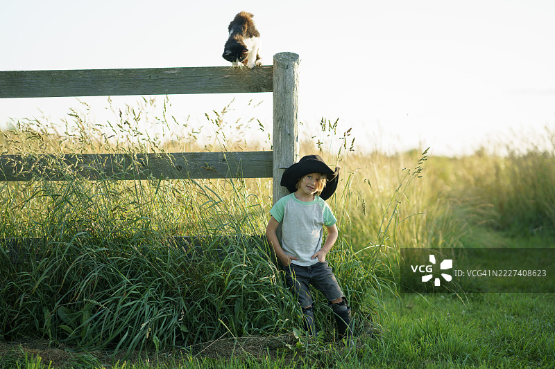 穿着黑帽子的孩子在乡村田野的围栏旁与猫咪相伴图片素材
