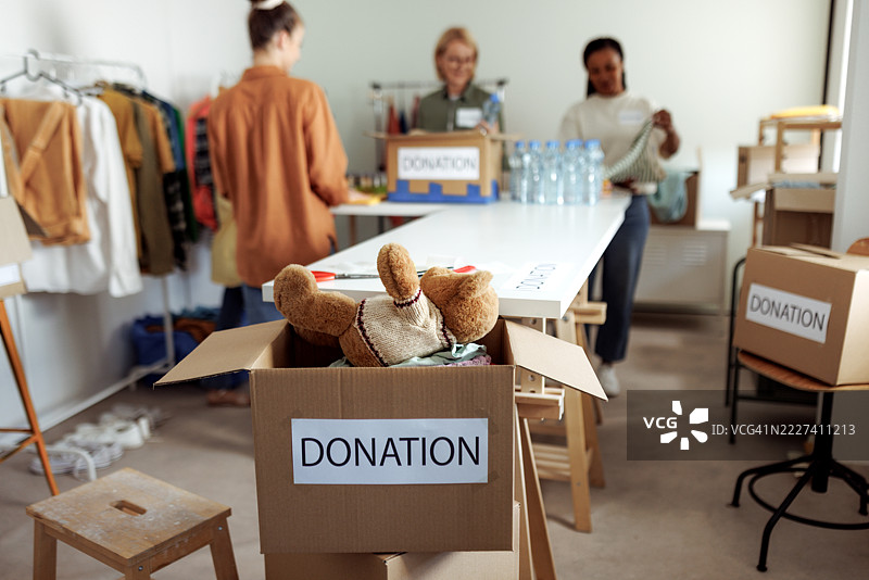 捐赠箱旁边有一只泰迪熊，背景中有女性正在打包物品。图片素材