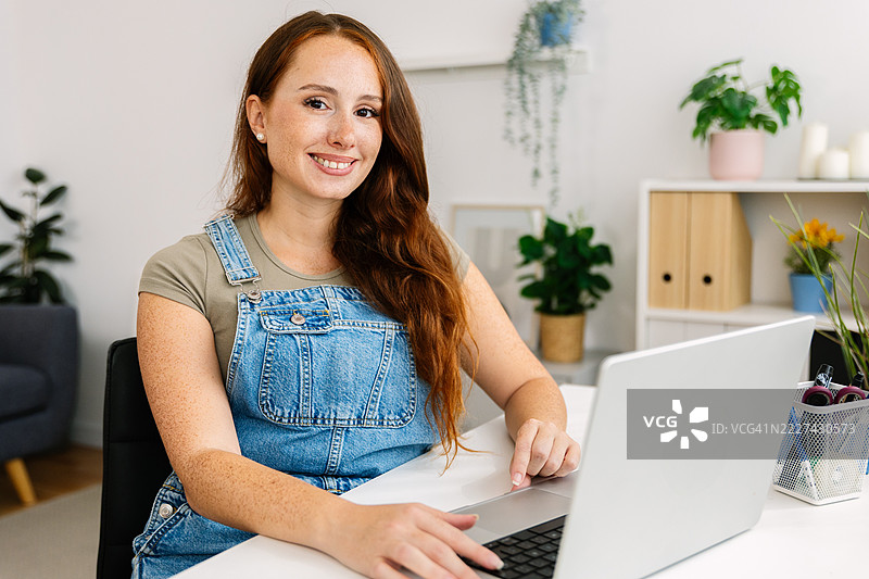 年轻快乐的千禧一代女性在家办公的工作场所微笑着看向镜头，坐在笔记本电脑前。图片素材