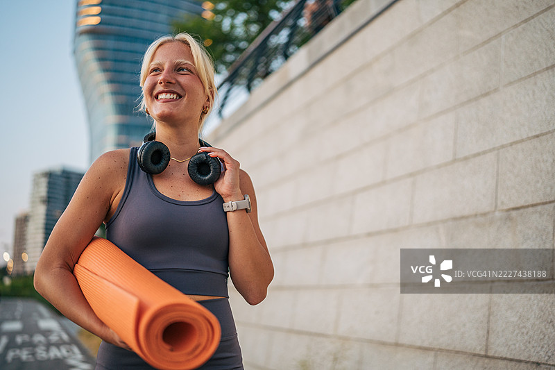 微笑的运动员在城市中走路，手里拿着瑜伽垫图片素材