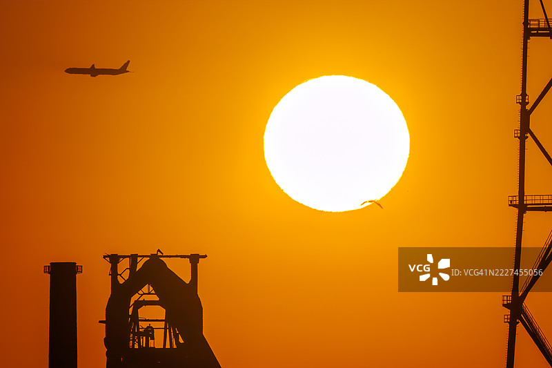 第一缕阳光与一架客机和鸟类的剪影。它即将降落在附近的羽田机场。第一缕阳光越过运河对岸的烟囱和电厂、钢铁厂。图片素材