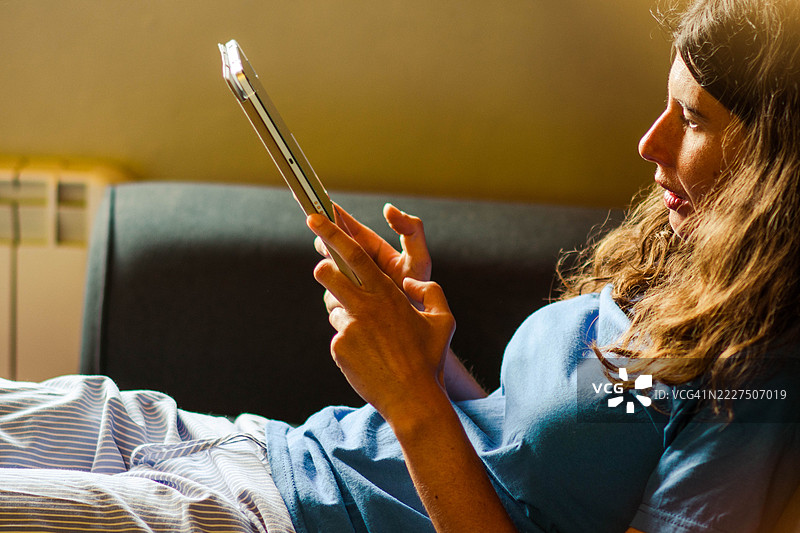 女人在家中舒适地躺在沙发上使用平板电脑。图片素材