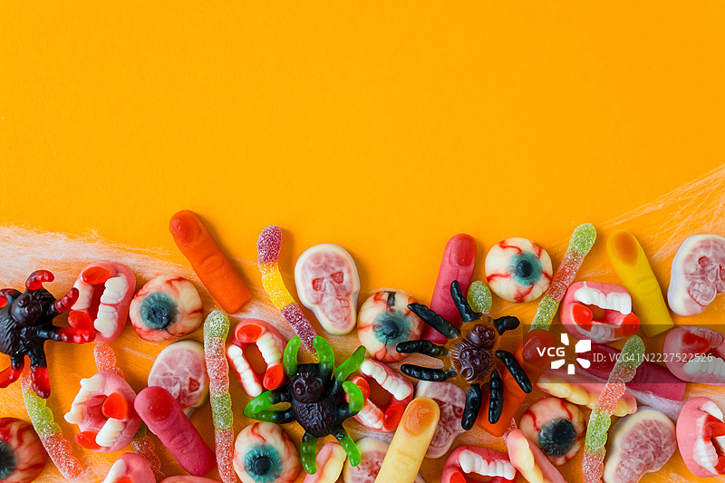 色彩鲜艳的万圣节主题软糖特写，包括软糖蜘蛛、眼球、獠牙、手指和骷髅，背景为橙色。图片素材
