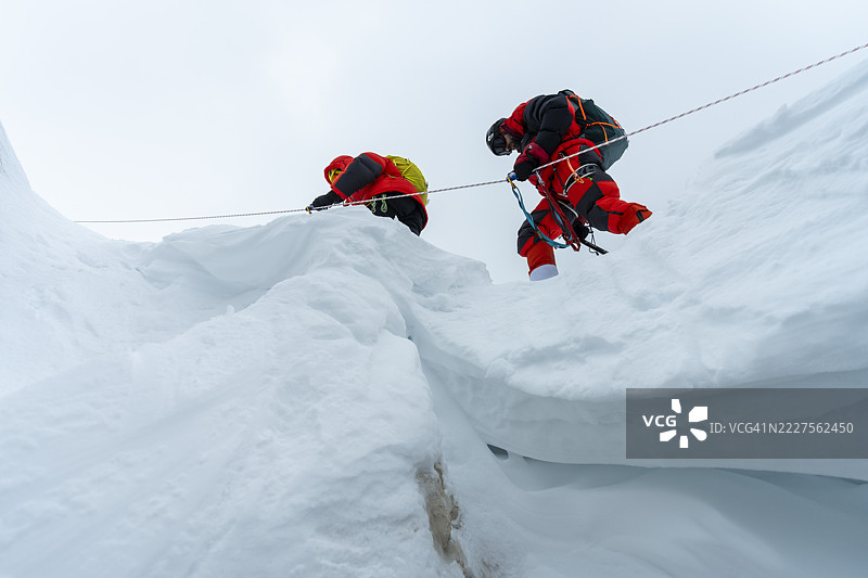 经验丰富的登山者穿着红色装备，携带绳索穿越雪裂缝，在阴云密布的天空下。图片素材
