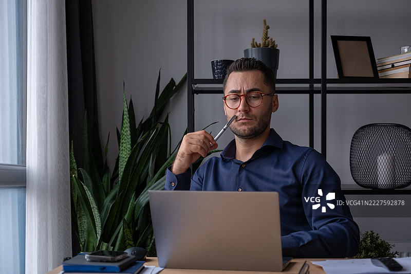 中年男子戴着眼镜，在现代办公室环境中思考并使用笔记本电脑工作图片素材
