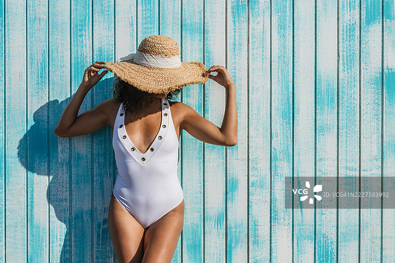 穿着泳衣和帽子的女人靠在蓝色木墙上，享受阳光明媚的日子图片素材