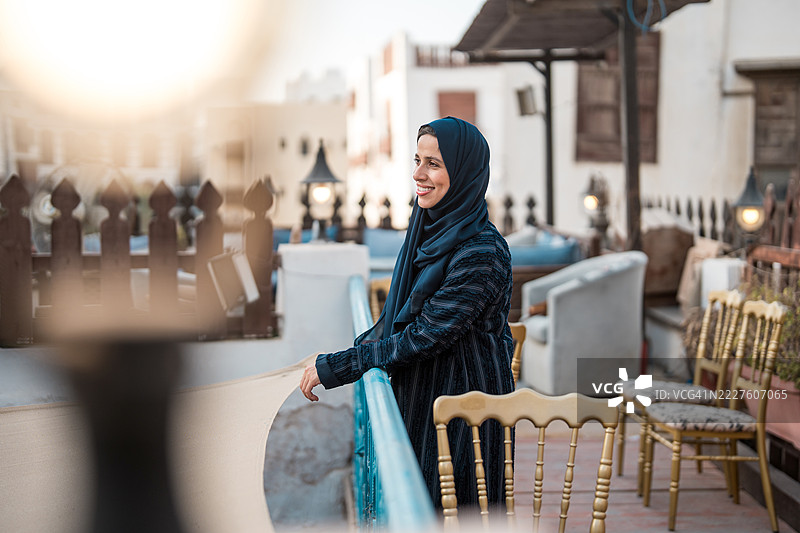 优雅的阿拉伯女性穿着传统黑色阿巴亚，在历史悠久的阿尔巴拉德屋顶露台上微笑图片素材