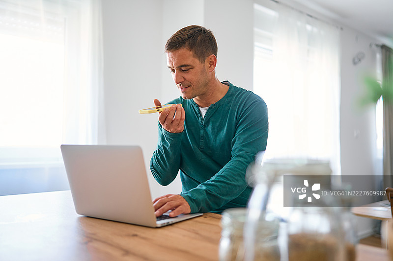 在家工作的男人使用手机上的语音识别软件并在笔记本电脑上打字图片素材