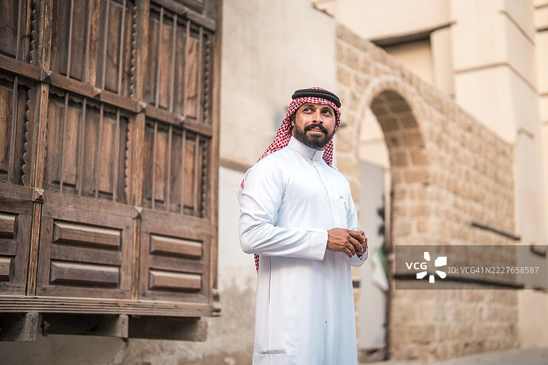 中年阿拉伯男子穿着传统白色长袍，站在阿尔巴拉德的历史建筑旁图片素材